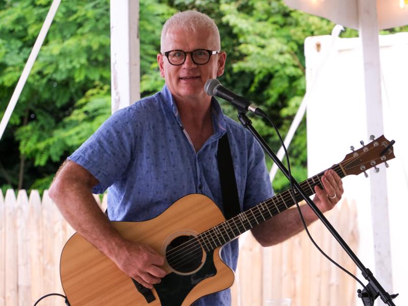 Steve Zaat smiling behind a microphone with his acoustic guitar at an outdoor gazebo performance