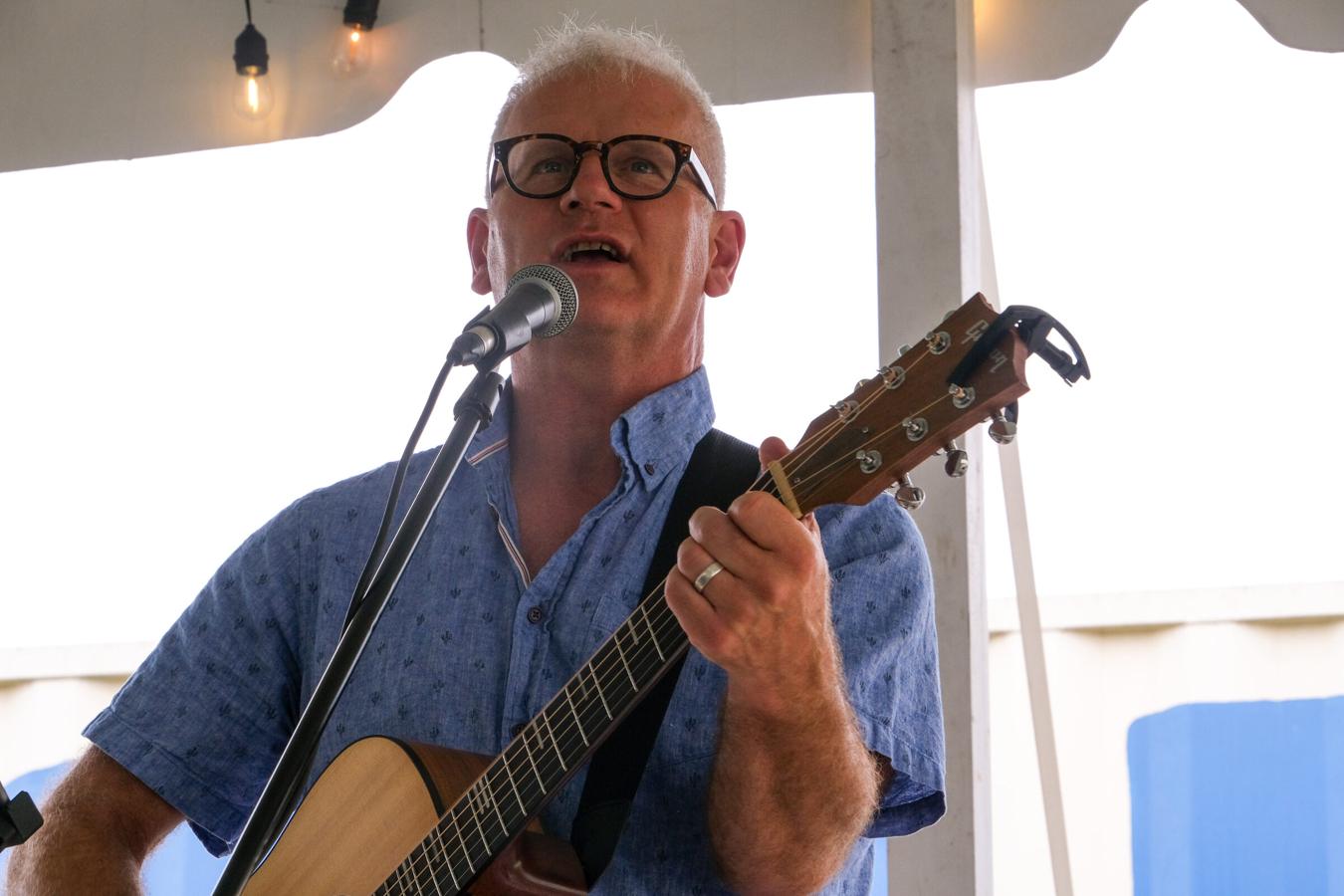 Steve Zaat singing into a microphone while playing acoustic guitar at an outdoor summer show