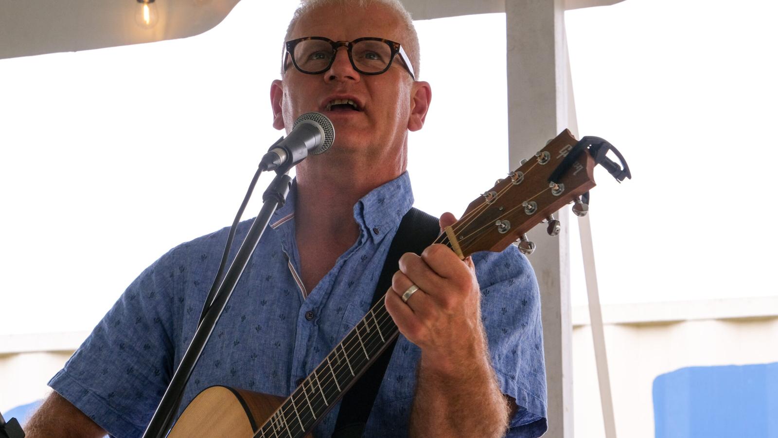 Steve Zaat singing into a microphone while playing acoustic guitar at an outdoor summer show