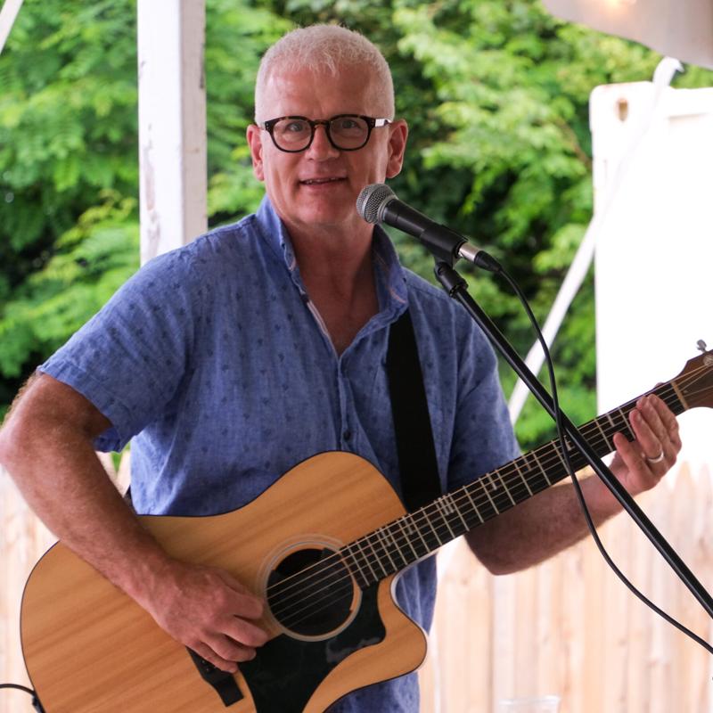 Steve Zaat smiling behind a microphone with his acoustic guitar at an outdoor gazebo performance